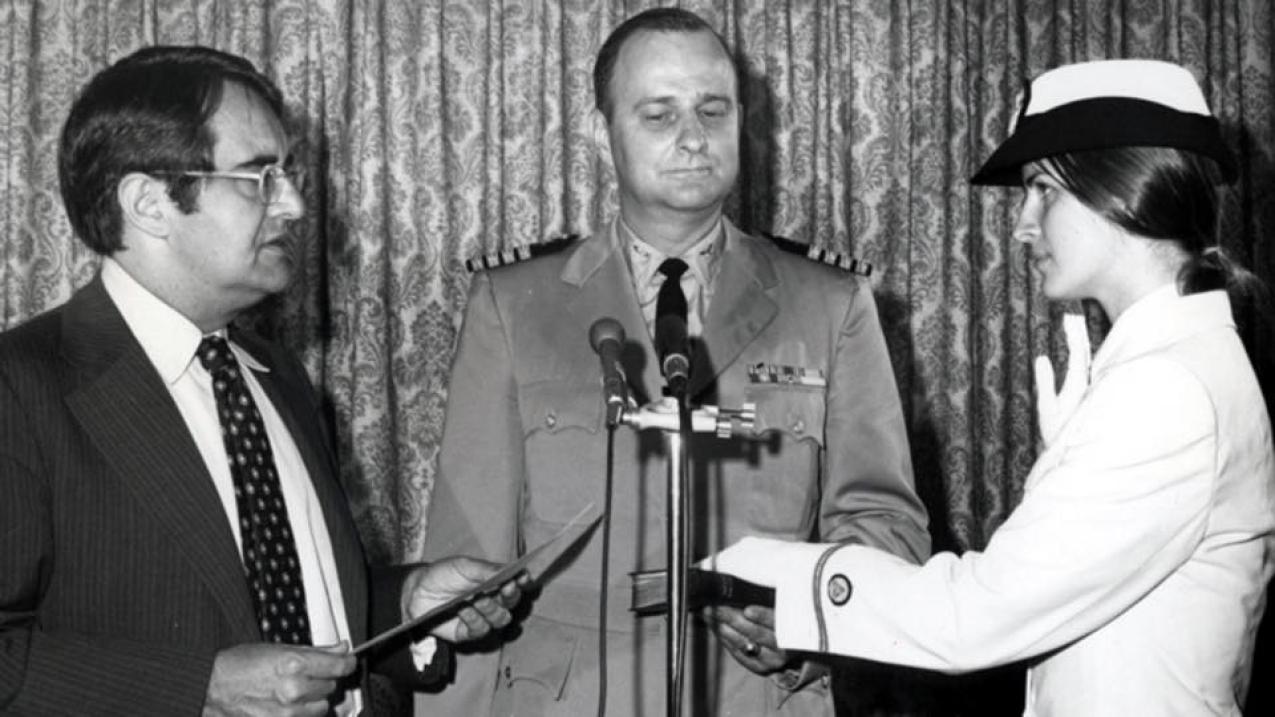 A black-and-white photograph of Ensign Pamela Chelgren was sworn in by Secretary of Commerce Peter G. Peterson on July 6, 1972. Her father, Navy Captain John Chelgren is in the center holding the Bible