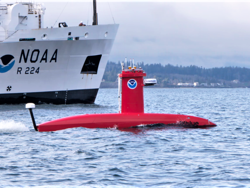 A bright orange submarine-shaped vehicle on the water with a NOAA ship behind it