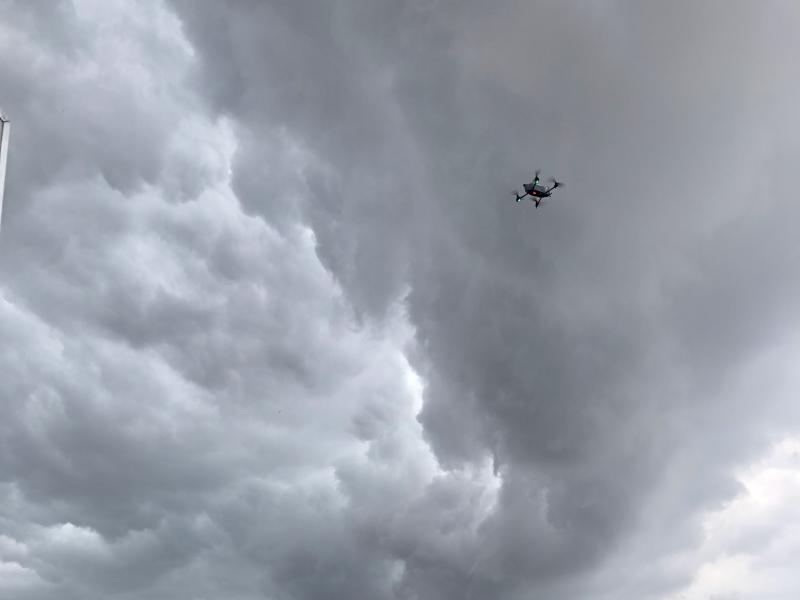 An uncrewed aircraft hovers in the air over a field with an orange landing pad. The sky is gray as though a storm is forming.