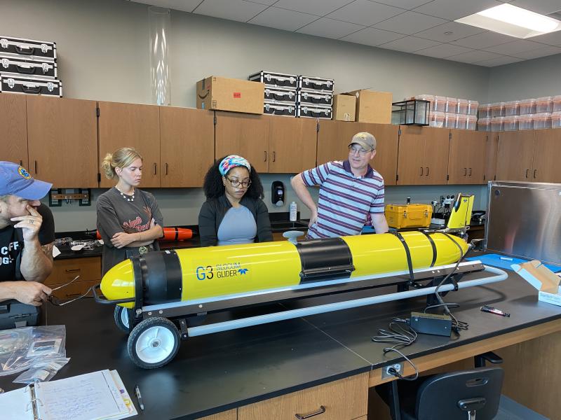 Knauss Fellow, Chesna Cox, with other glider pilots around her, programs a yellow Slocum glider sitting on a table in front of them.