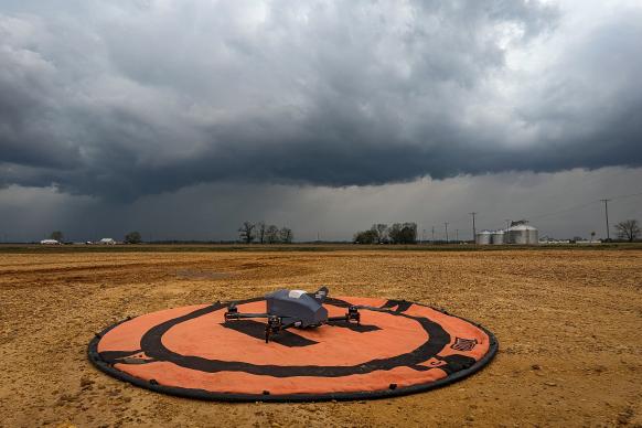 A gray uncrewed aircraft sits on a orange launch pad in an open field. A storm with dark clouds forms in the distance