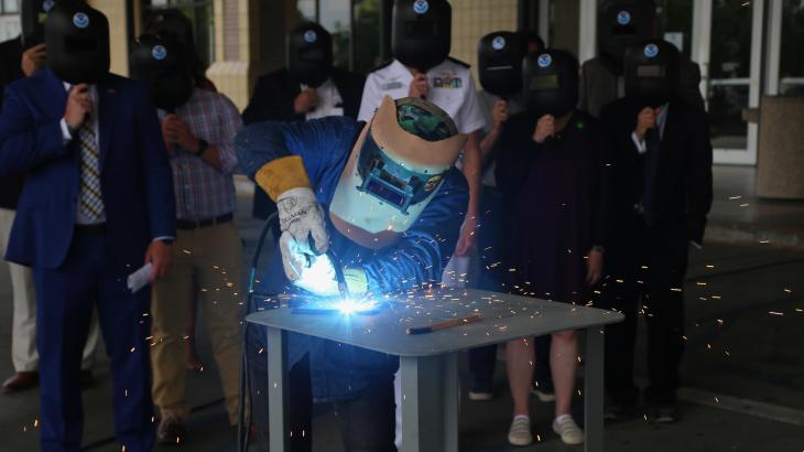 A welder working on a metal plate while a group of people look on