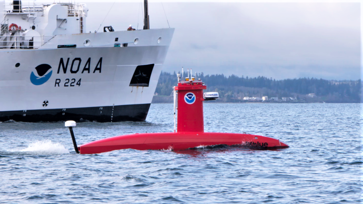 A bright orange submarine-shaped vehicle on the water with a NOAA ship behind it