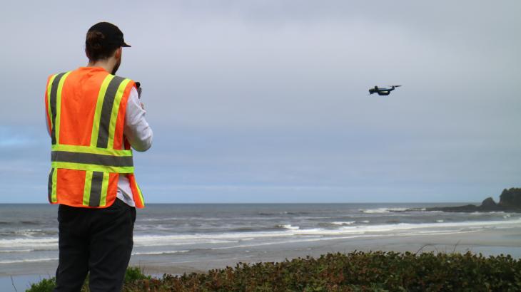 A man in an orange and yellow safety vest stands on a beach with his back to the camera. He is piloting a small drone that is flying in the distance over the sand.