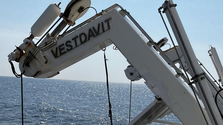 A gray davit arm extends over the edge of a ship. The DriX deployment gondola with a red DriX are being lowered by the davit.