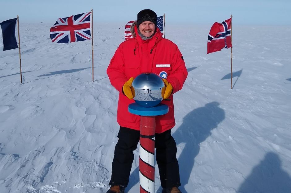 A NOAA Corps officer in a bright orange jacket at the striped South Pole marker