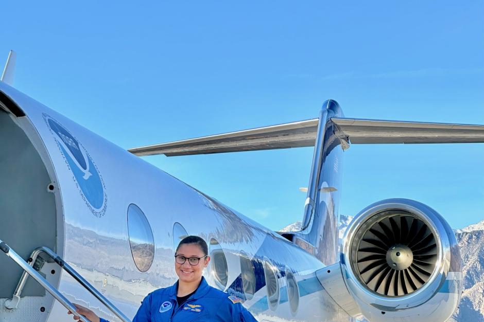 A NOAA Corps officer in a blue flightsuit standing on the stairs to a NOAA jet