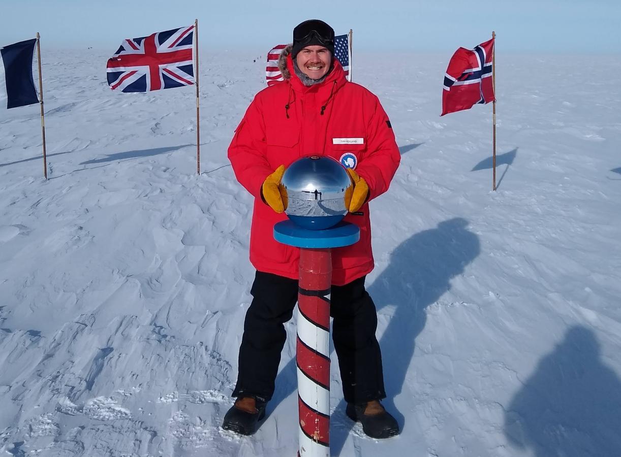 A NOAA Corps officer in a bright orange jacket at the striped South Pole marker