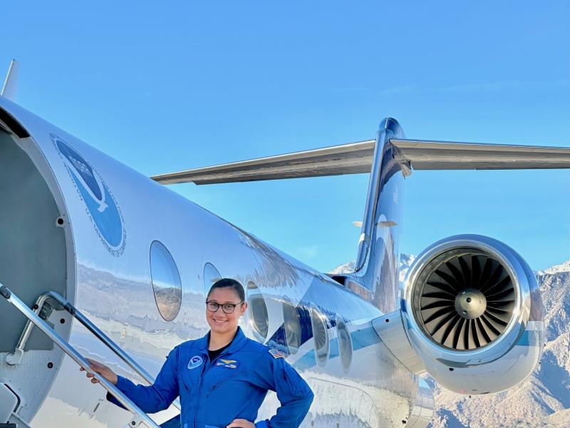 A NOAA Corps officer in a blue flightsuit standing on the stairs to a NOAA jet