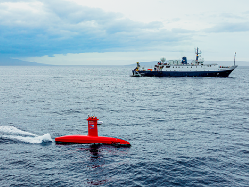 A red uncrewed marine system transits on top of the ocean with the E/V Nautilus in the background.