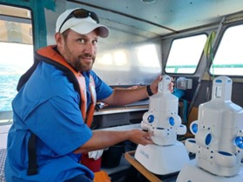 A NOAA scientist poses for the camera in a blue sun shirt, white hat, and orange life jacket while in the cabin of a boat. They are holding a small, white uncrewed marine systems called the Hydrus.