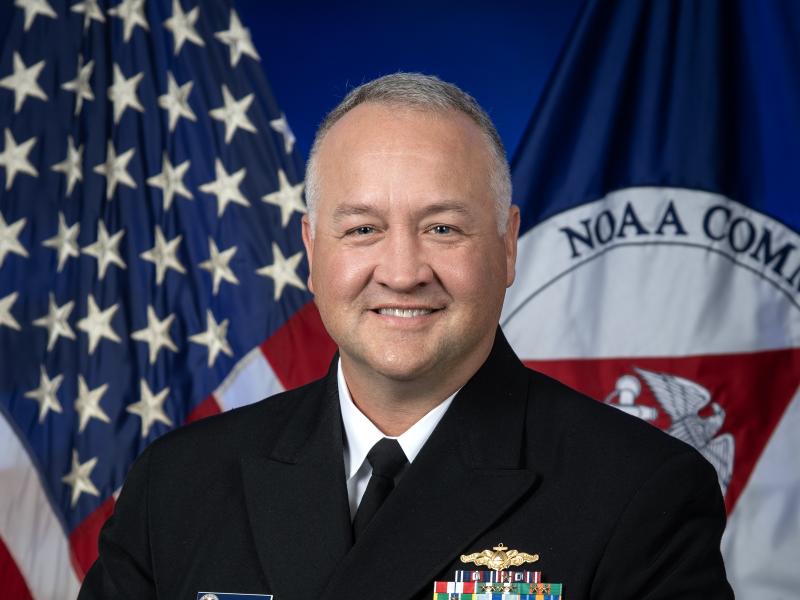 Rear Admiral Chad Cary in dress blue uniform with U.S. and NOAA Corps flags behind him