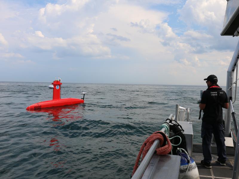 An operator stands on the side of a small boat as it pilots a red DriX uncrewed surface vehicle in the distance.