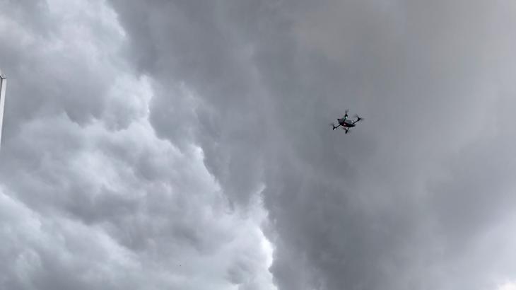 An uncrewed aircraft hovers in the air over a field with an orange landing pad. The sky is gray as though a storm is forming.