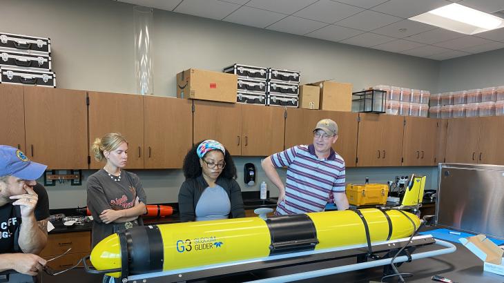 Knauss Fellow, Chesna Cox, with other glider pilots around her, programs a yellow Slocum glider sitting on a table in front of them.