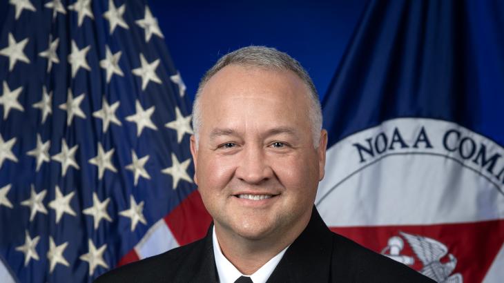 Rear Admiral Chad Cary in dress blue uniform with U.S. and NOAA Corps flags behind him