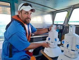 A NOAA scientist poses for the camera in a blue sun shirt, white hat, and orange life jacket while in the cabin of a boat. They are holding a small, white uncrewed marine systems called the Hydrus.