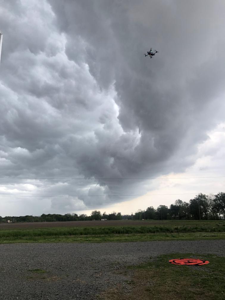 An uncrewed aircraft hovers in the air over a field with an orange landing pad. The sky is gray as though a storm is forming.