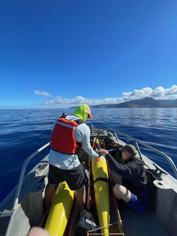 Two people hold on to yellow underwater gliders in the front of a small boat as they approach an island.